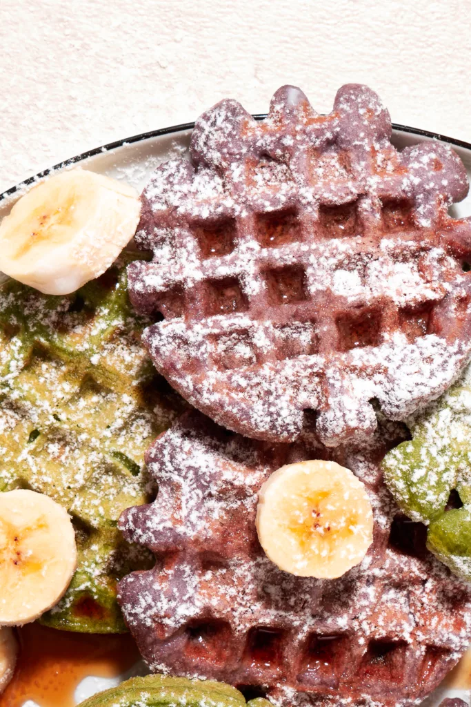 ube and matcha mochi waffles drizzled with syrup and topped with bananas and powdered sugar