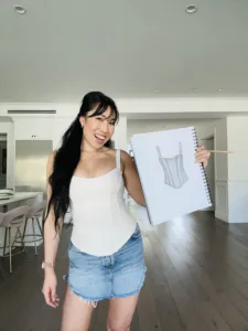 Cassey wearing the Coconut Cream Corset Cami corset tank top with a jean skirt, while holding the design sketch to the product. 